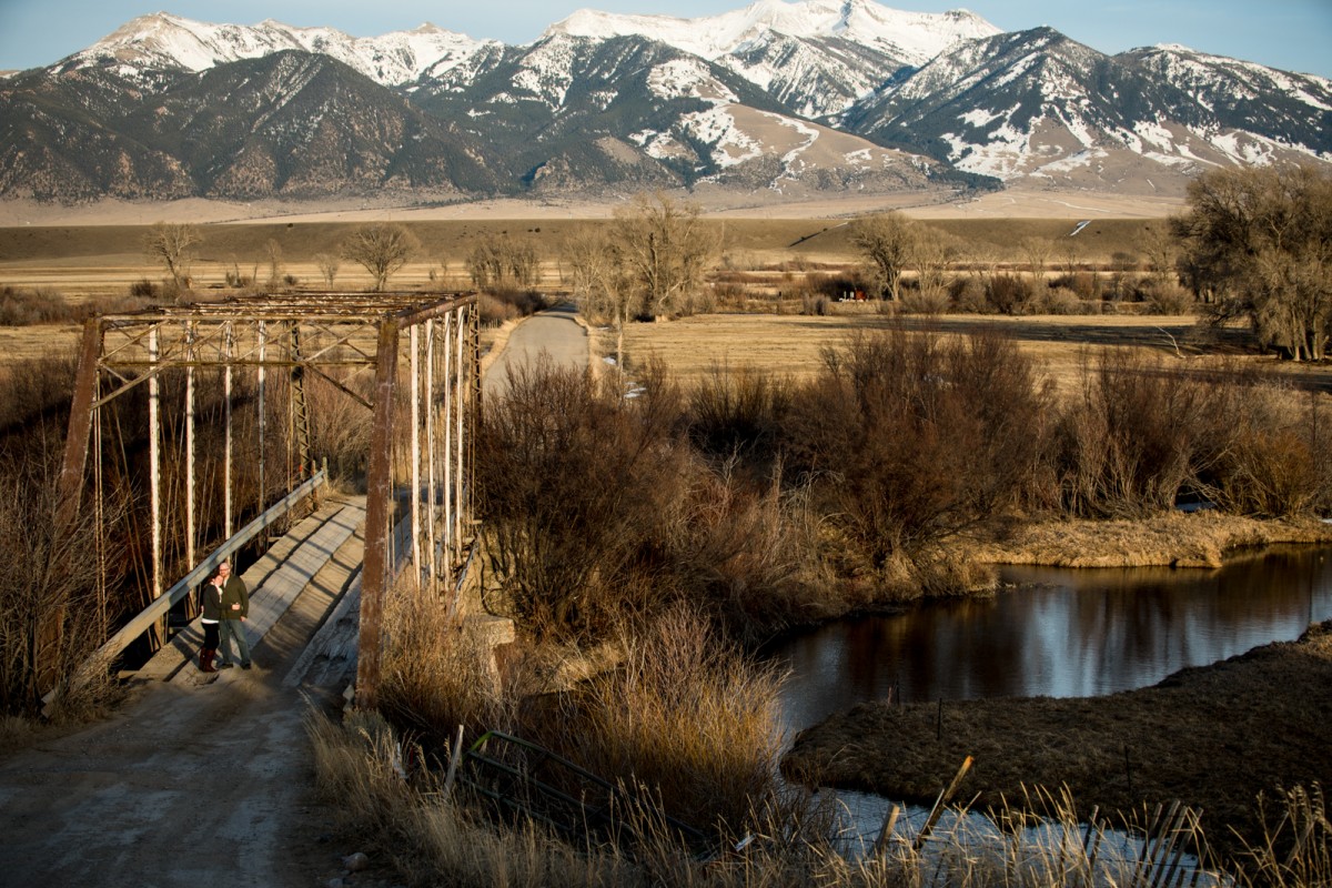 couple-standing-on-bridge-with-mountain-backdrop-in-Ennis-Montana-Engagement-Photos