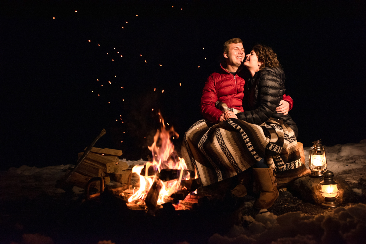 engaged-couple-laugh-together-around-campfire-during-Hyalite-stargazing-engagement-portraits