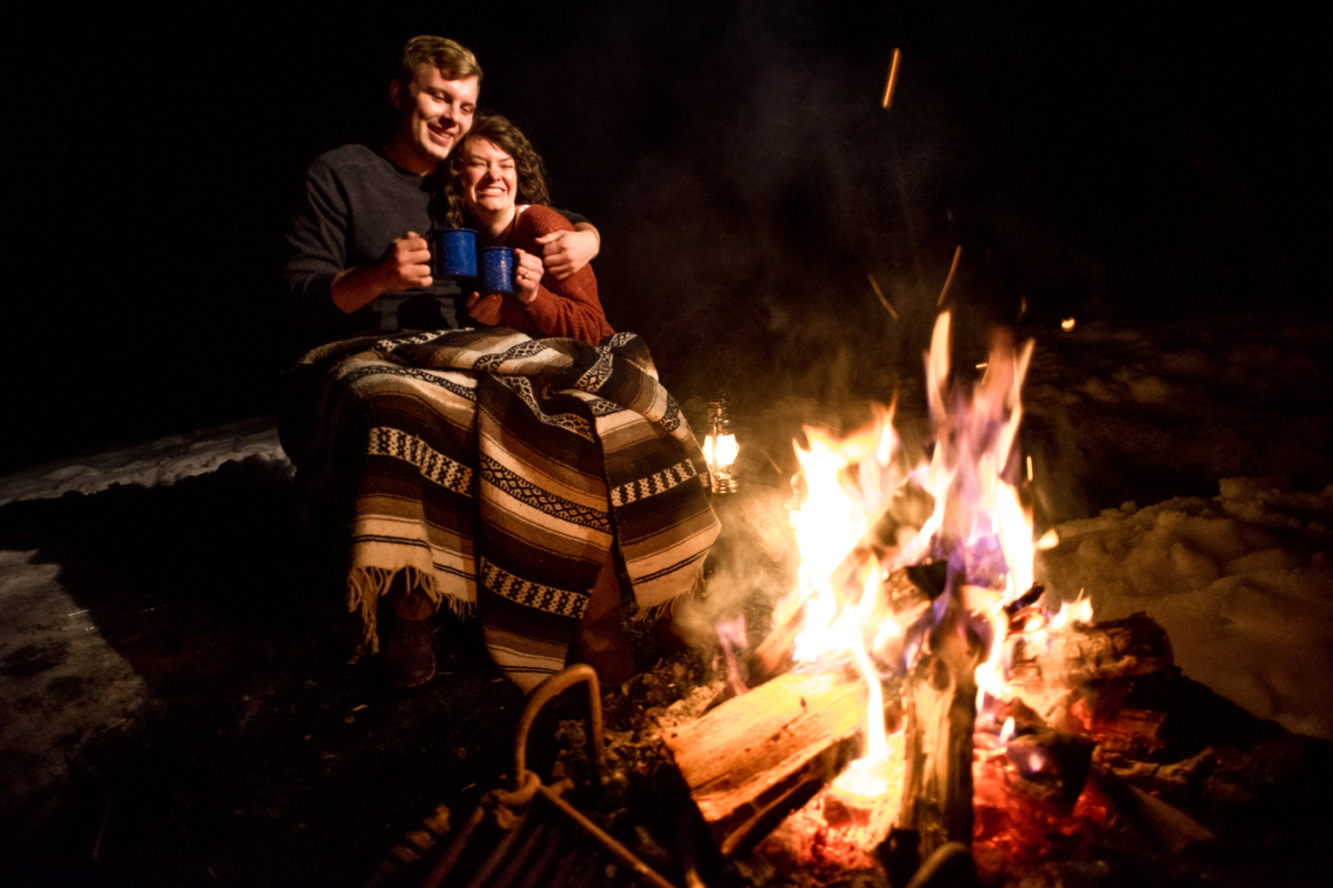 engaged-couple-drink-hot-chocolate-around-campfire-during-Hyalite-stargazing-engagement-portraits