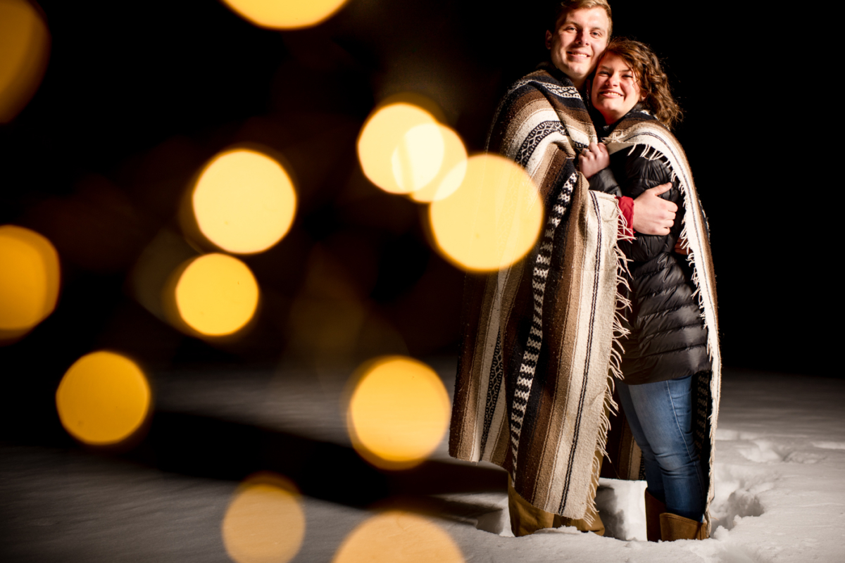 engaged-couple-stand-on-reservoir-under-golden-lights-during-Hyalite-stargazing-engagement