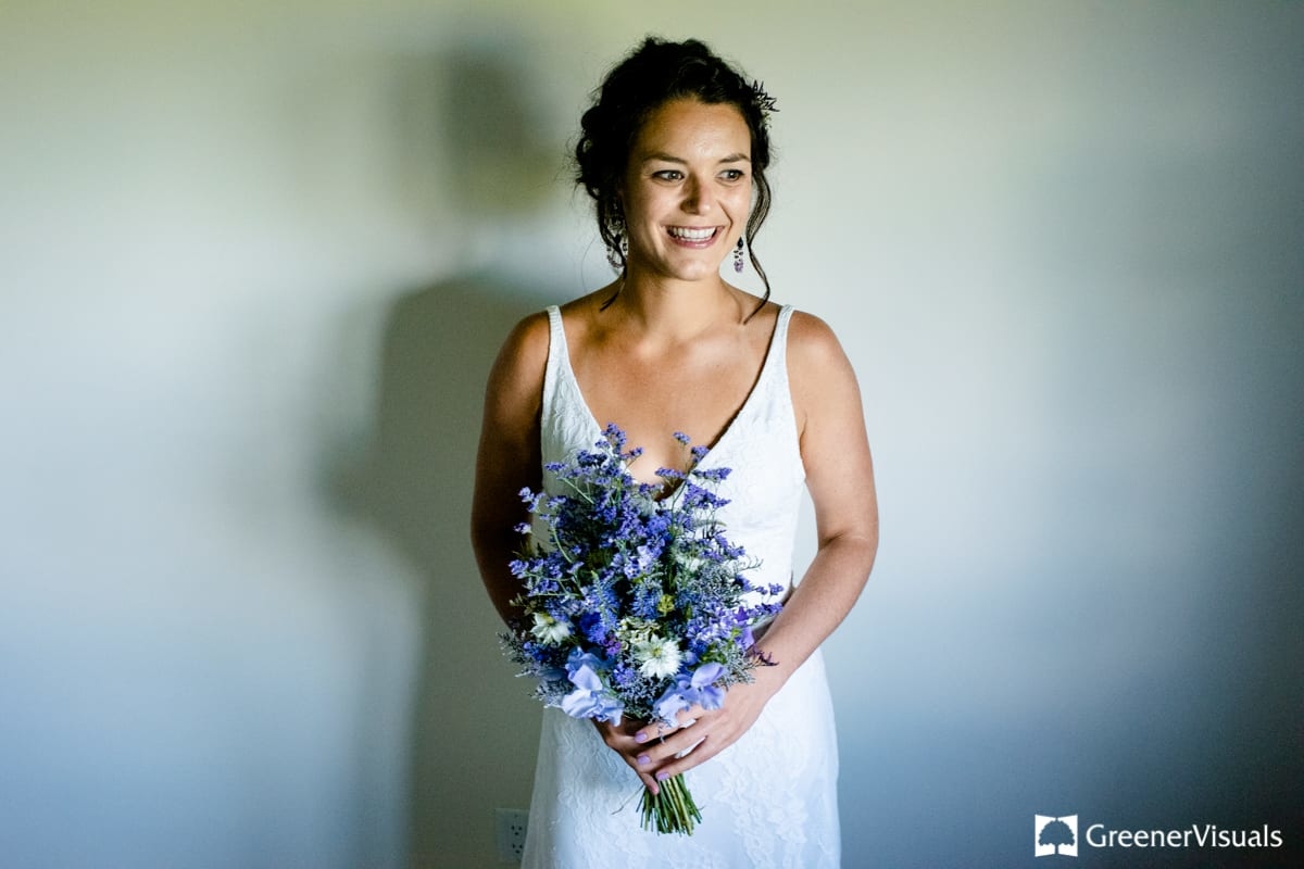 bride-stands-with-blue-flowers-against-white-wall-during-Countryside-Bozeman-Montana-Wedding