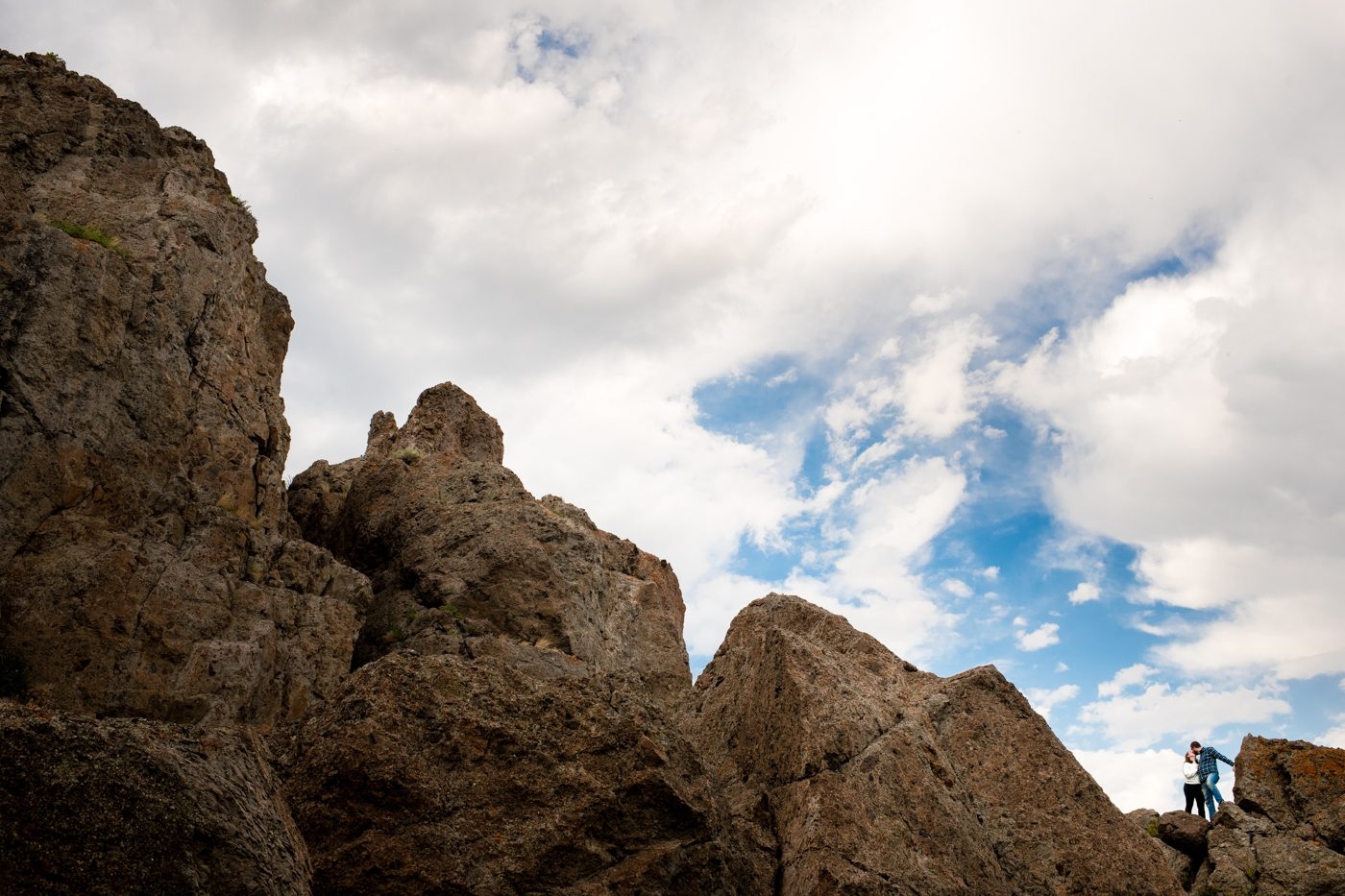 Engaged-couple-stand-on-dramatic-rocks-against-blue-sky