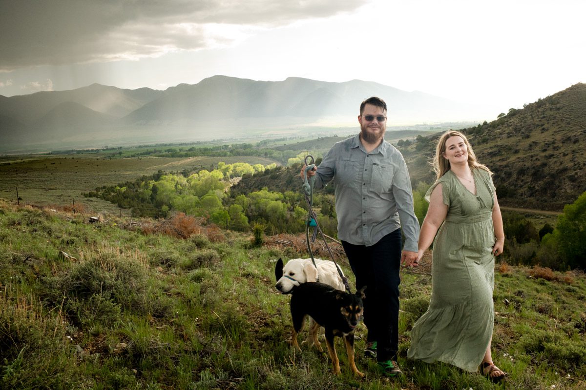 engaged-couple-walk-with-dogs-on-green-mountainside-during-Montana-Ruby-Valley